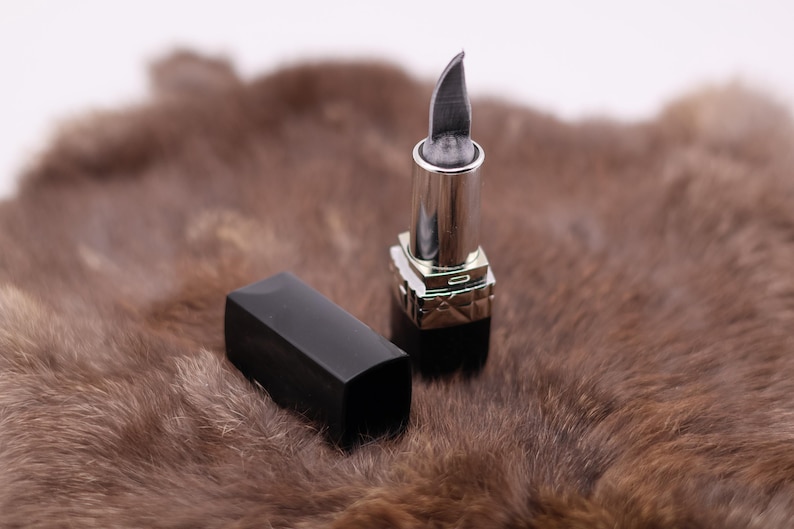 Puede incluir: Un pintalabios plateado con una punta afilada, sobre una superficie de piel marr&oacute;n. El pintalabios est&aacute; cerrado y tiene una tapa negra.