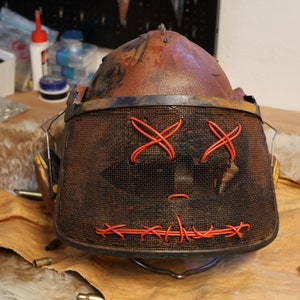 Könnte beinhalten: Ein rostiger Metallhelm mit einem Drahtgitter-Gesichtsmask. Die Maske hat rote Nähte, die ein X für die Augen und eine Linie für den Mund bilden.