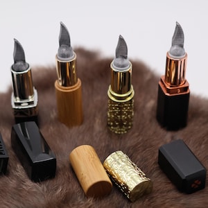 Könnte beinhalten: Acht verschiedene Lippenstift-Röhrchen mit verschiedenen Oberflächen, darunter Schwarz, Gold und Holz. Die Röhrchen sind in einer Reihe auf einer braunen, pelzigen Oberfläche angeordnet.