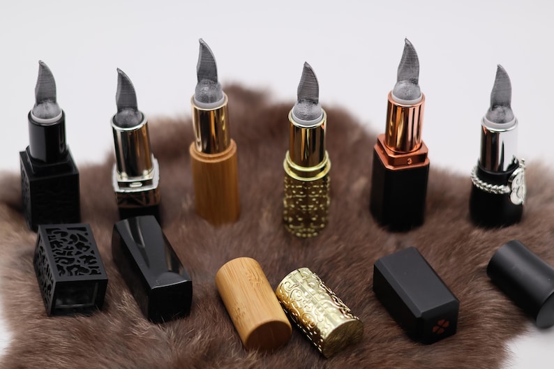 Puede incluir: Ocho tubos de l&aacute;piz labial diferentes con varios acabados, incluyendo negro, dorado y madera. Los tubos est&aacute;n dispuestos en una fila sobre una superficie peluda marr&oacute;n.