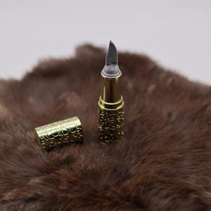 Puede incluir: Un tubo de l&aacute;piz labial de color dorado con una cuchilla plateada que se extiende desde la parte superior. El tubo est&aacute; sobre una superficie de piel marr&oacute;n.