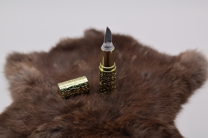 Puede incluir: Un tubo de l&aacute;piz labial de color dorado con una cuchilla plateada que se extiende desde la parte superior. El tubo est&aacute; sobre una superficie de piel marr&oacute;n.