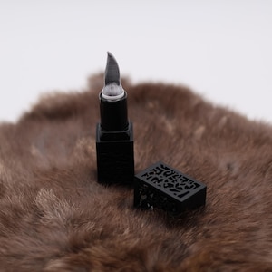 Puede incluir: Barra de labios negra con un dise&ntilde;o decorativo e intrincado en el estuche. La barra de labios est&aacute; sobre una superficie de piel marr&oacute;n.