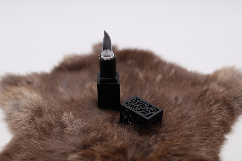 Puede incluir: Barra de labios negra con un dise&ntilde;o decorativo e intrincado en el estuche. La barra de labios est&aacute; sobre una superficie de piel marr&oacute;n.