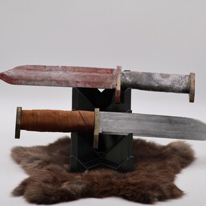 Könnte beinhalten: Zwei Messer mit Metallklingen und Holzgriffen werden auf einem schwarzen Ständer über einem braunen Fell ausgestellt. Das obere Messer hat eine rote Klinge und einen grauen Griff. Das untere Messer hat eine silberne Klinge und einen braunen Griff.