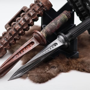 Può includere: Tre coltelli diversi con design unici. I coltelli sono esposti su un supporto nero con uno sfondo di pelliccia marrone. I coltelli hanno una varietà di finiture, tra cui nero, marrone e argento.