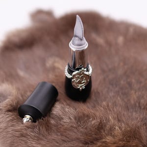 Puede incluir: Dos tubos de l&aacute;piz labial negros con detalles plateados. Un tubo tiene una tapa plateada puntiaguda y el otro tiene una tapa plateada redondeada. Los tubos est&aacute;n sobre una superficie de piel marr&oacute;n.