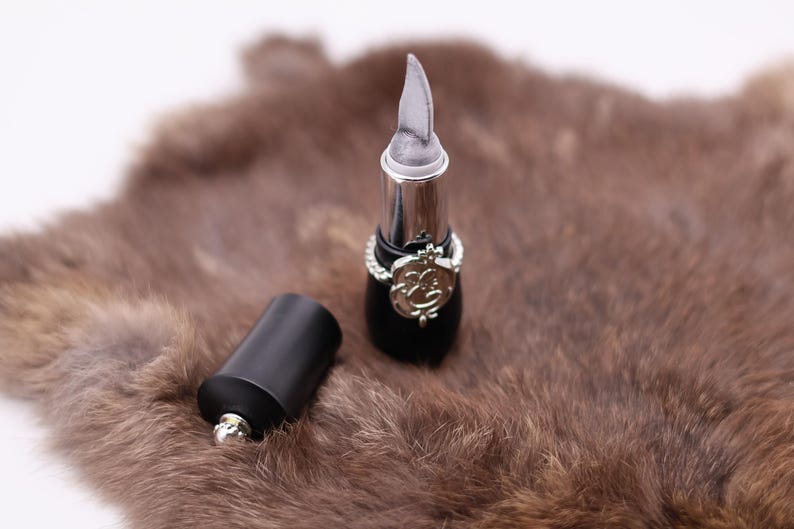 Puede incluir: Dos tubos de l&aacute;piz labial negros con detalles plateados. Un tubo tiene una tapa plateada puntiaguda y el otro tiene una tapa plateada redondeada. Los tubos est&aacute;n sobre una superficie de piel marr&oacute;n.