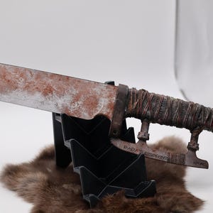 Könnte beinhalten: Ein rostiges, verwittertes Metallsäbel mit einer braunen Lederwicklung am Griff. Das Säbel ist auf einem schwarzen Ständer mit dem Text "DADA'S EISBRUCH" auf der Klinge ausgestellt.