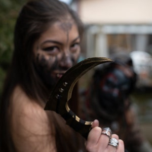 以下が含まれることがあります： 黒髪で顔に黒いペイントをした女性が、湾曲した黒と金のナイフを持っている。彼女は指に銀の指輪をしている。