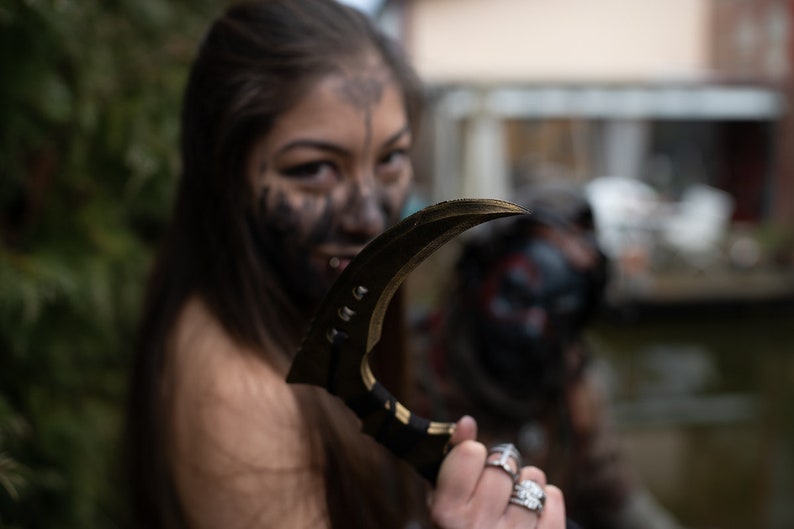 以下が含まれることがあります： 黒髪で顔に黒いペイントをした女性が、湾曲した黒と金のナイフを持っている。彼女は指に銀の指輪をしている。