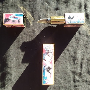 Könnte beinhalten: Drei goldene Lippenstift-Röhrchen mit bunten Designs. Die Lippenstift-Röhrchen zeigen ein Schwarzweißbild einer Frau mit einer Krone auf dem Kopf, einem schwarzen Vogel und einem bunten abstrakten Design.