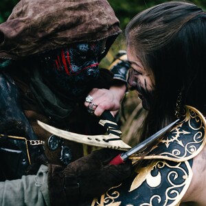 以下が含まれることがあります： フード付きの茶色のマントを着て、赤い模様のついた黒いフェイスマスクをしている人と、黒と金の装飾的な肩当てをしている人のクローズアップ。彼らは手にナイフを持っている。
