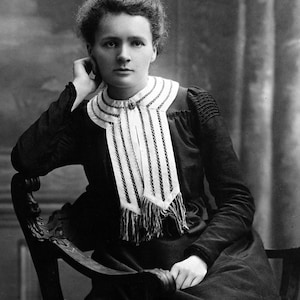 May include: A black and white portrait of a woman with dark hair styled in a bun. She is wearing a dark dress with a white and black striped collar. She is sitting in a chair with her hand resting on her chin.