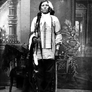 May include: A black and white portrait of a Native American man wearing traditional clothing and a beaded necklace. He is standing in front of a painted backdrop.