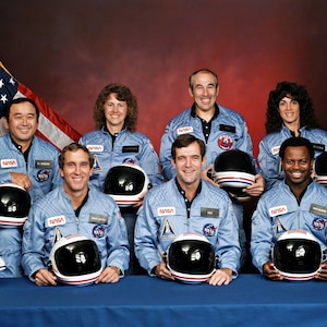 May include: A group of eight astronauts in blue flight suits stand in front of an American flag. They are holding helmets and a model of the space shuttle. The astronauts are smiling and looking at the camera.