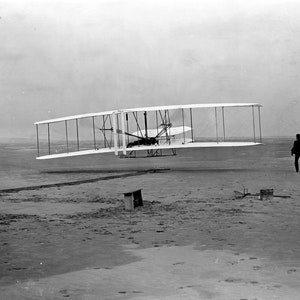 May include: A black and white photograph of the Wright brothers' first successful airplane, the Wright Flyer, taking off from the sand at Kitty Hawk, North Carolina. A man in a dark suit stands on the beach watching the plane take off.