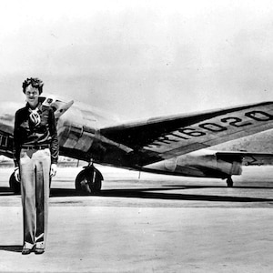May include: A black and white photo of a woman standing in front of a silver airplane. The airplane has the number "NR16020" on the tail.