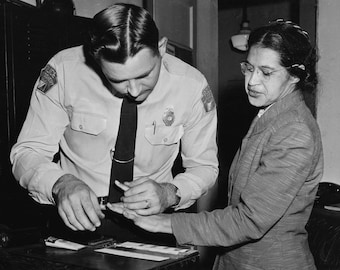 May include: A black and white photo of a police officer taking a woman's fingerprints. The woman is wearing a suit jacket and glasses. The officer is wearing a white shirt and tie.