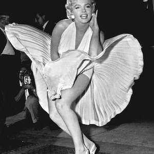 May include: A black and white photo of a woman in a white dress with the skirt blowing up in the wind. She is smiling and holding her hand to her face.