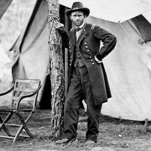 May include: A black and white photograph of a man in a military uniform standing next to a tree. He is wearing a hat and has a serious expression on his face.