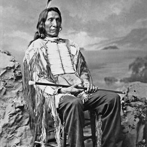 May include: A black and white portrait of a Native American man wearing traditional clothing and a feathered headdress. He is sitting on a chair and holding a walking stick.