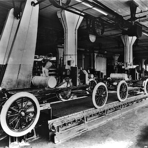 May include: Black and white photo of a car factory assembly line.  The photo shows several car chassis on a moving conveyor belt.  The chassis are being assembled in a factory setting.