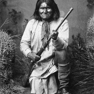 May include: Black and white portrait of a Native American man kneeling, holding a rifle. He wears a light-colored shirt, a scarf, and boots. The background features foliage and a textured backdrop.