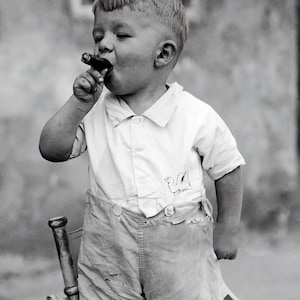 May include: A young boy in a white button-down shirt and shorts is holding a cigar in his mouth. He is looking to the right and has a serious expression on his face.