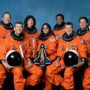 May include: A group of seven astronauts in orange space suits stand in front of an American flag and an Israeli flag. They are smiling and looking at the camera. The astronaut in the center is holding a mission patch that says "STS-107".