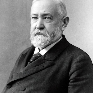 May include: A black and white portrait of a man with a full white beard and mustache. He is wearing a dark suit with a white shirt and a patterned tie.