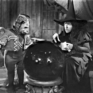 May include: Black and white image of a scene from a movie featuring a witch in a tall hat and a winged monkey. The witch is holding cards and looking at a large crystal ball. The monkey is standing next to the ball, wearing a costume.
