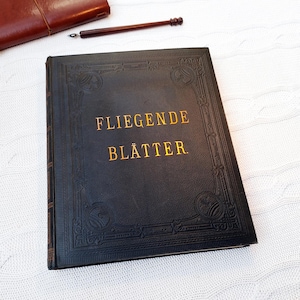 Puede incluir: Un libro negro vintage con letras doradas que dice "FLIEGENDE BLÄTTER". El libro tiene una cubierta en relieve con bordes decorativos. Un diario de cuero marrón y un bolígrafo están en el fondo.