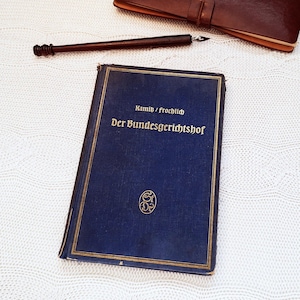 Könnte beinhalten: Ein altes Buch mit dunkelblauem Einband und goldener Schrift, betitelt "Der Bundesgerichtshof". Ein brauner Stift und ein ledergebundenes Notizbuch sind ebenfalls zu sehen. Das Buch liegt auf einer weißen, strukturierten Oberfläche.
