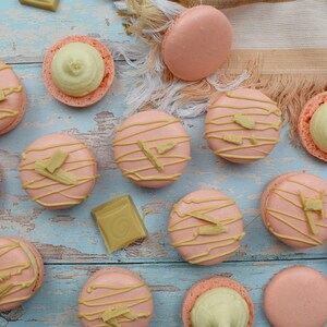 May include: A collection of pink macarons with white chocolate drizzled on top and white chocolate pieces on a blue wooden background.