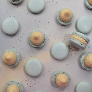 May include: A collection of pastel blue and light yellow macarons arranged on a light gray surface. The macarons are decorated with a swirl of light yellow frosting.