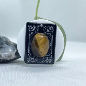 May include: A rectangular pendant with a black background and white decorative edges. The pendant features an oval tiger's eye stone in the center. The pendant is hanging on a white surface.