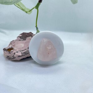 Könnte beinhalten: Ein polierter, hellrosa Rosenquarz-Kristall liegt in einer kleinen, weißen Keramikschale. Links von der Schale befindet sich ein Stück rosa und braunes Gestein. Im Hintergrund sind ein grüner Stiel und Blätter zu sehen.