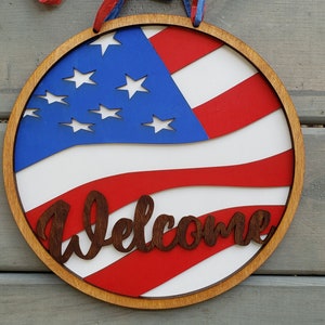 Puede incluir: Un letrero redondo de madera con un diseño de bandera estadounidense roja, blanca y azul. El letrero tiene la palabra "Welcome" escrita en letras marrones.