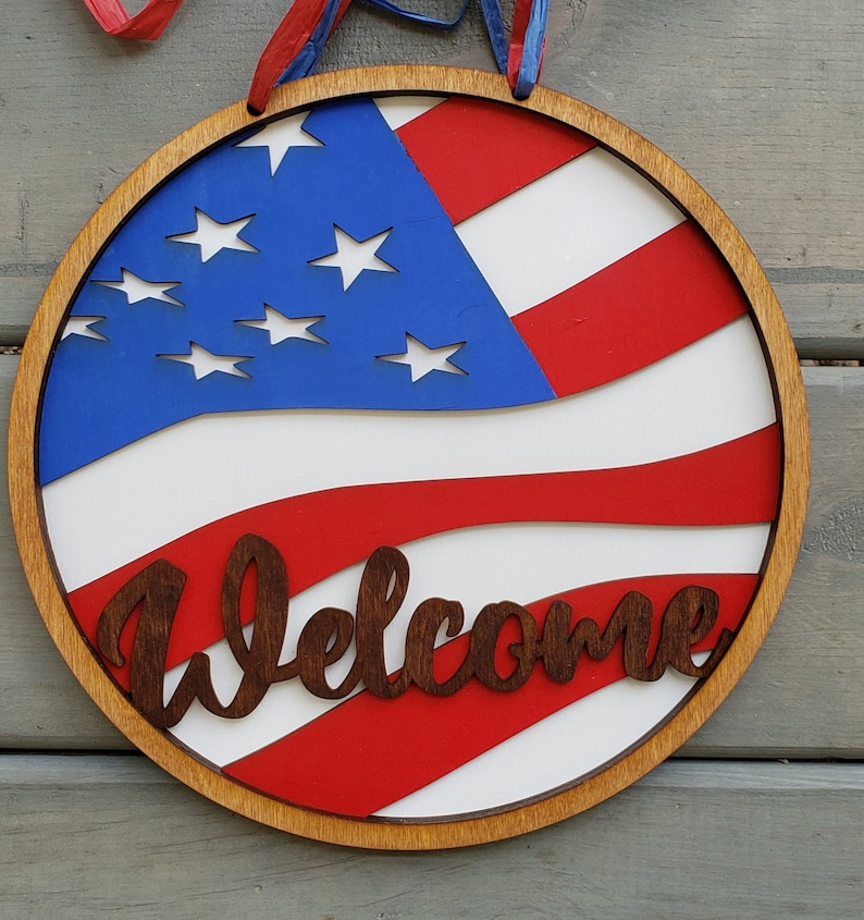Op de afbeelding: Een rond houten bord met een rood, wit en blauw Amerikaans vlagontwerp. Het bord heeft de tekst "Welcome" in bruine letters.