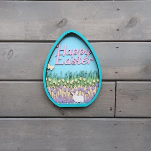 May include: A blue and white wooden egg-shaped ornament with the words "Happy Easter" in pink lettering. The ornament features a white bunny rabbit, green grass, and yellow and purple flowers.