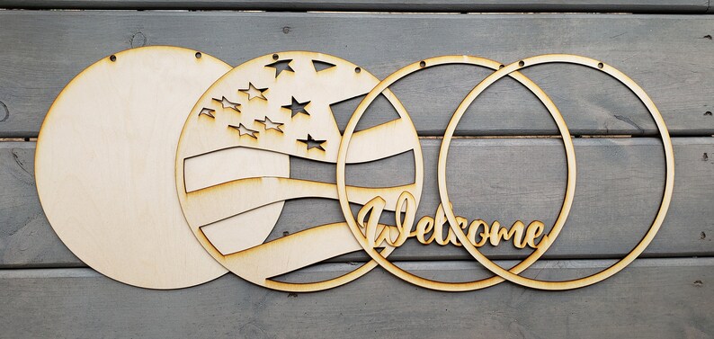 Op de afbeelding: Drie onafgewerkte houten cirkels met uitsparingen. De eerste cirkel is leeg. De tweede cirkel heeft een uitsparing van de Amerikaanse vlag. De derde cirkel heeft het woord "Welkom" uitgesneden.