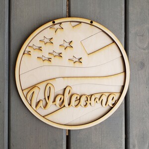 Op de afbeelding: Een rond houten welkom-bord met een Amerikaans vlagontwerp in rood, wit en blauw. Het bord heeft een gat bovenaan om het op te hangen.