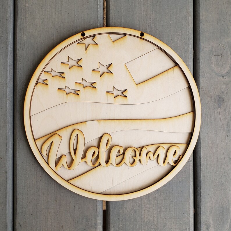 Op de afbeelding: Een rond houten welkom-bord met een Amerikaans vlagontwerp in rood, wit en blauw. Het bord heeft een gat bovenaan om het op te hangen.