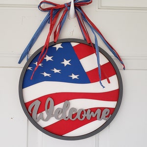 Op de afbeelding: Een ronde houten deurhanger met een rood, wit en blauw Amerikaans vlagontwerp en het woord "Welkom" in zilveren letters. De deurhanger is versierd met rode en blauwe linten.