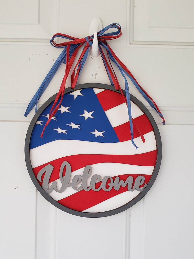 Op de afbeelding: Een ronde houten deurhanger met een rood, wit en blauw Amerikaans vlagontwerp en het woord "Welkom" in zilveren letters. De deurhanger is versierd met rode en blauwe linten.