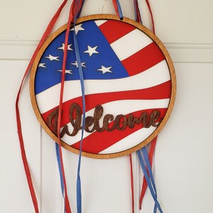 Op de afbeelding: Een rond houten welkomsbord met een Amerikaans vlagontwerp en het woord "Welkom" in een bruin lettertype. Het bord is versierd met rode en blauwe linten.