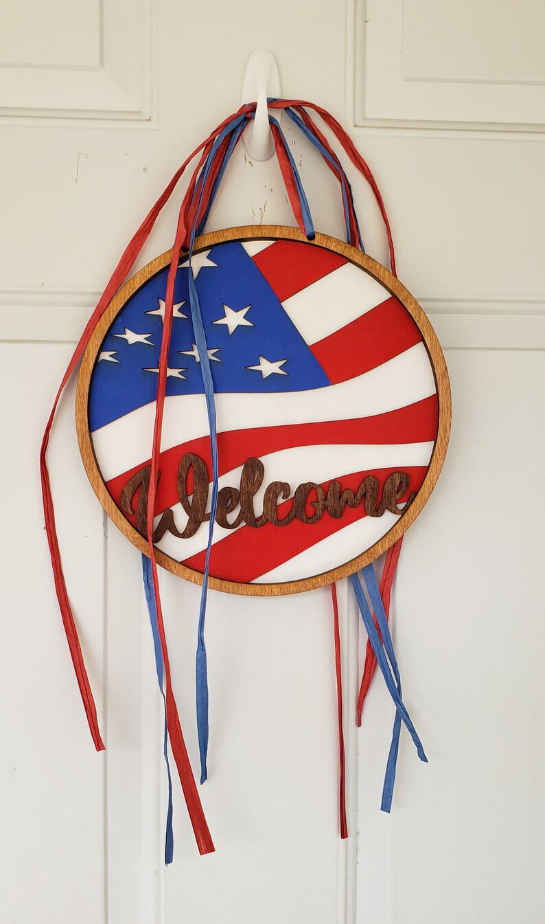 Op de afbeelding: Een rond houten welkomsbord met een Amerikaans vlagontwerp en het woord "Welkom" in een bruin lettertype. Het bord is versierd met rode en blauwe linten.