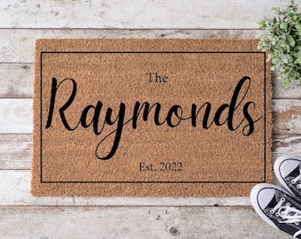 May include: A brown coir doormat with a black border and the text "The Raymonds Est. 2022" in black cursive font.