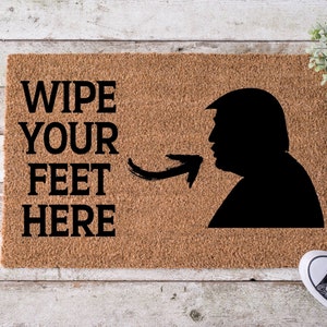 Op de afbeelding: Een bruine kokosvezel deurmat met de tekst "Wipe Your Feet Here" in zwarte letters. Aan de rechterkant van de mat is een zwart silhouet van een hoofd en schouders van een persoon, met een pijl die naar de tekst wijst.